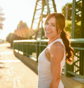A woman in a white tank top stands confidently on a sunlit bridge, with a lush green backdrop and a clear blue sky A woman in a white tank top stands confidently on a sunlit bridge, with a lush green backdrop and a clear blue sky